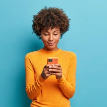 Calm serious dark skinned young woman plays games on phone or sends text messages connected to high speed internet uses modern technologies dressed in casual jumper poses against blue background. Brasil é o país com mais ataques a dispositivos móveis na América Latina