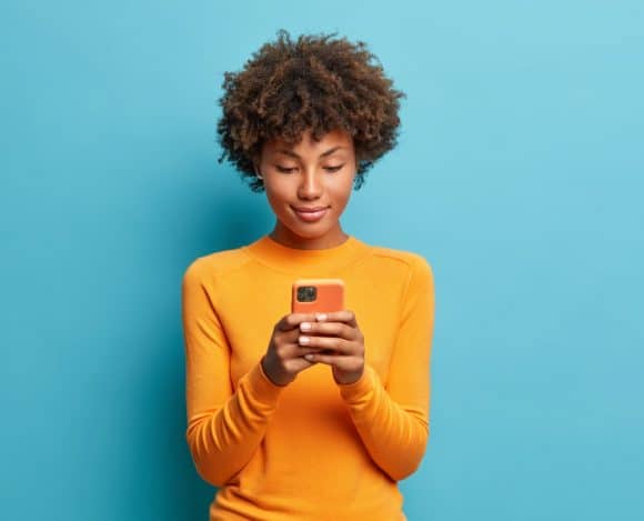 Calm serious dark skinned young woman plays games on phone or sends text messages connected to high speed internet uses modern technologies dressed in casual jumper poses against blue background. Brasil é o país com mais ataques a dispositivos móveis na América Latina